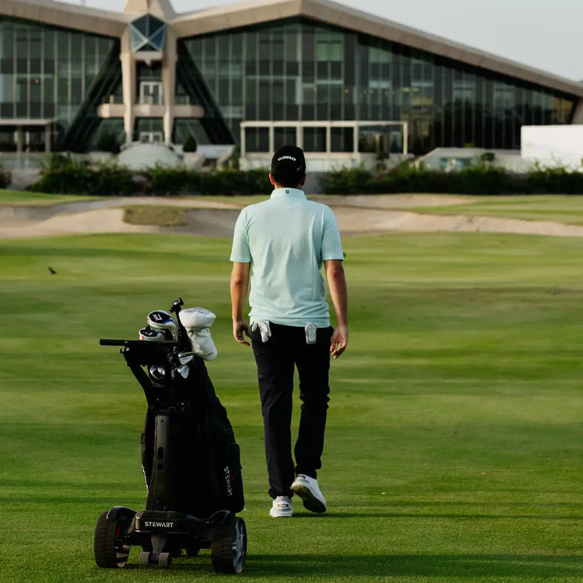 Man walking on a golf course with a golf bag cart, building in the background