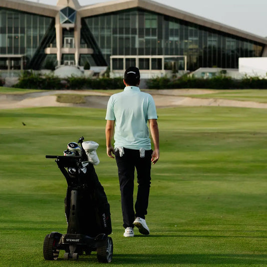 Man walking on a golf course with a golf bag cart, building in the background